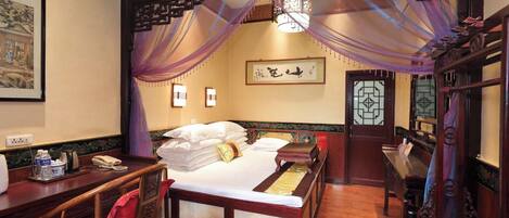 A traditional room with a bed, a desk, and a chair. There is a decorative screen above the bed and a wall-mounted light fixture.