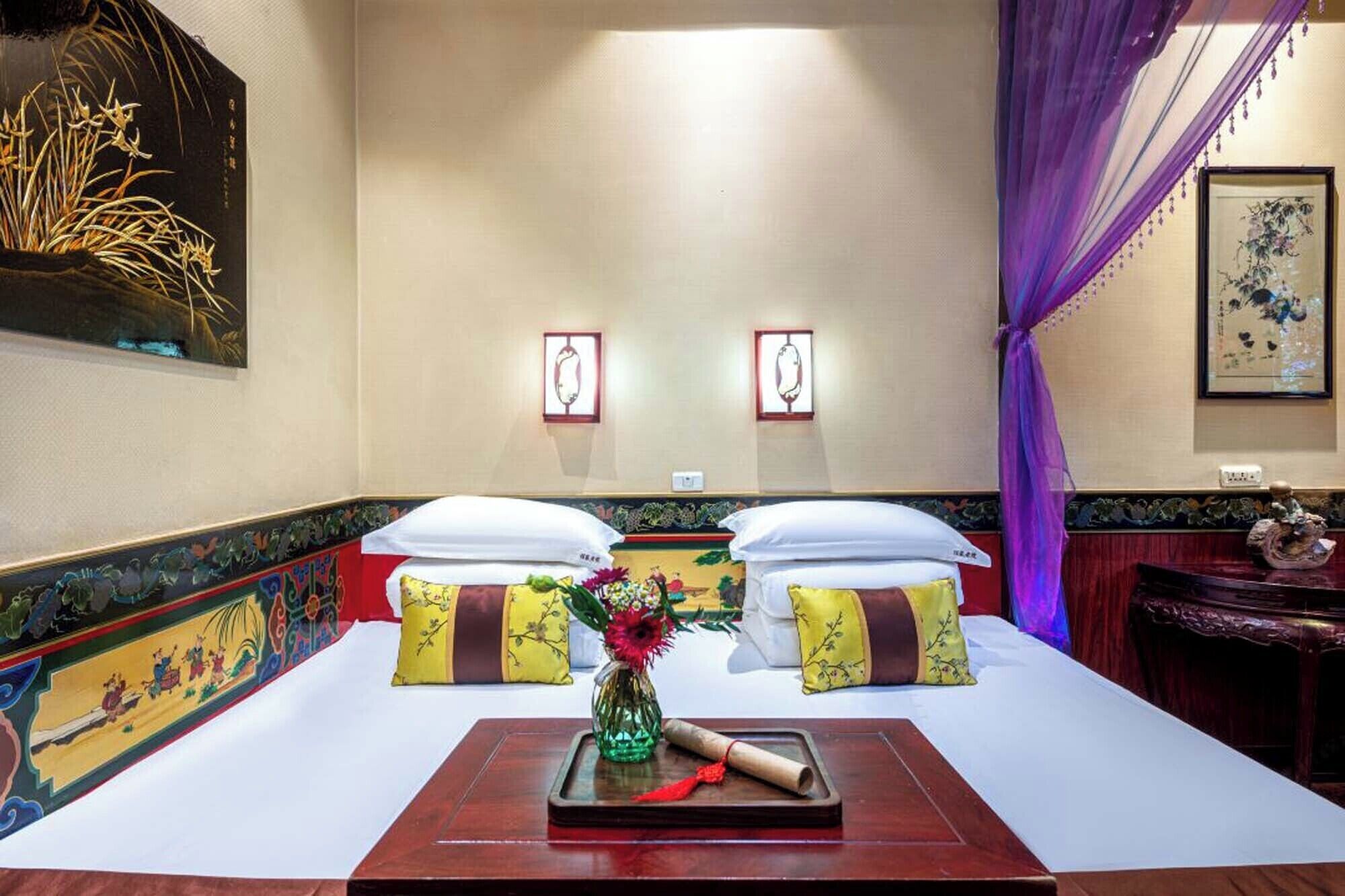 A hotel room with two beds, each with a pillow and a vase of flowers. There are wall-mounted picture frames and a decorative vase on a side table.