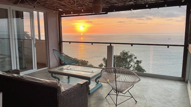 Un balcon avec une table en verre, deux chaises et un canapé en osier, donnant sur la mer au coucher du soleil.