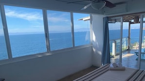 Une chambre avec une grande fenêtre donnant sur la mer, un lit recouvert d’une couverture à rayures et un ventilateur de plafond.