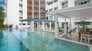 En swimmingpool med et overdækket område og udendørs siddepladser, omgivet af en bygning med flere etager og altaner.