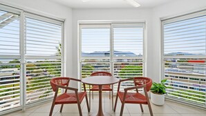 Ein Balkon mit rundem Tisch und vier Stühlen, große Fenster mit Jalousien und Blick auf eine Stadtlandschaft und Berge.