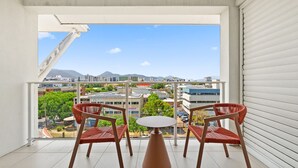 A balcony with two red wicker chairs and a small round table, overlooking a cityscape with buildings and trees.