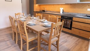Ein Essbereich mit einem Holztisch, gedeckt für vier Personen, umgeben von Holzstühlen. Im Hintergrund ist eine Küche mit Holzausbau und Geschirrspüler zu sehen.