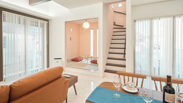 A modern living room with a sofa, dining table set for two, and a staircase leading to an upper floor.