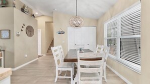 A dining area with a wooden table and white chairs, a chandelier, and a window with blinds.