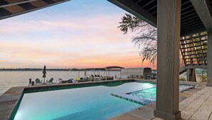 An infinity pool with a view of the sunset over the water, wooden deck, and lounge chairs.
