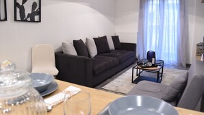 Ein modernes Wohnzimmer mit einem dunklen Sofa, einem weißen Sessel und einem Couchtisch. An der Wand hängen gerahmte Bilder und ein Fenster mit transparenten Vorhängen.