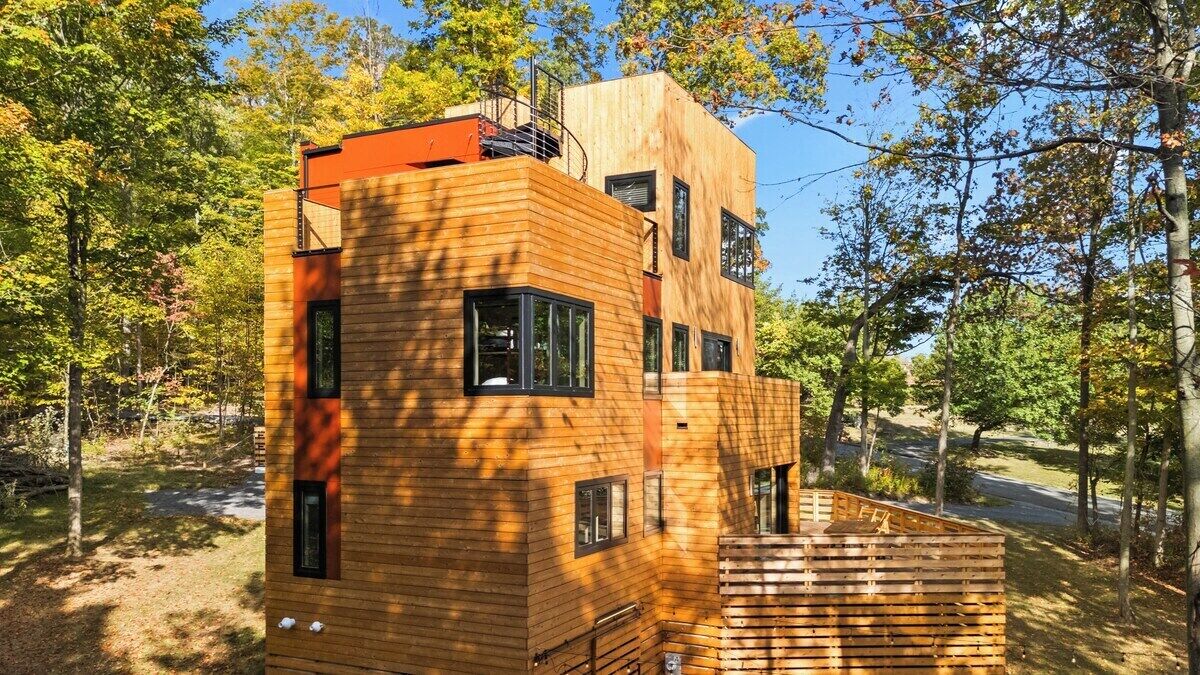 Une cabane en bois moderne, dotée de nombreuses fenêtres et d’une petite terrasse, entourée d’arbres.
