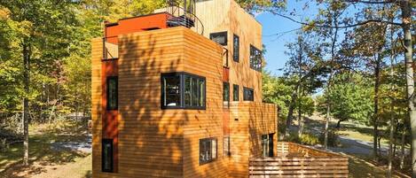 Une cabane en bois moderne, dotée de nombreuses fenêtres et d’une petite terrasse, entourée d’arbres.