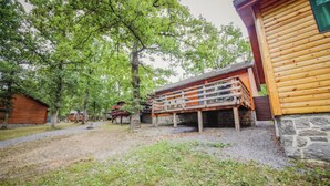A rustic cabin with a wooden deck and stone foundation, surrounded by trees and a gravel path.