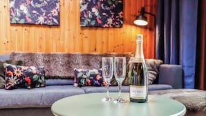 A room with a wooden wall, a sofa with floral cushions, a round table with a bottle and two glasses.