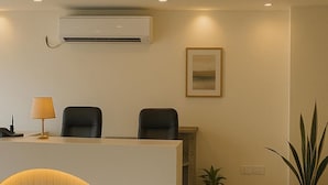 A reception area with a white reception desk, two black chairs, a lamp, an air conditioner, a framed picture on the wall, and two potted plants. - Hotel Maski Suites (Hyderabad)