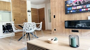 Ett modernt vardagsrum med ett matbord i trä, vita stolar, en tv som är monterad på väggen och ett köksområde med träskåp.
