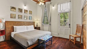 Un dormitorio con cabecera de madera, cama con ropa de cama blanca, banco de madera, ventilador de techo y ventana con cortinas.