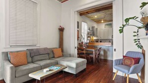 Una sala de estar con un sofá gris, una mesa de centro de madera y un sillón azul. Se ve una cocina al fondo.