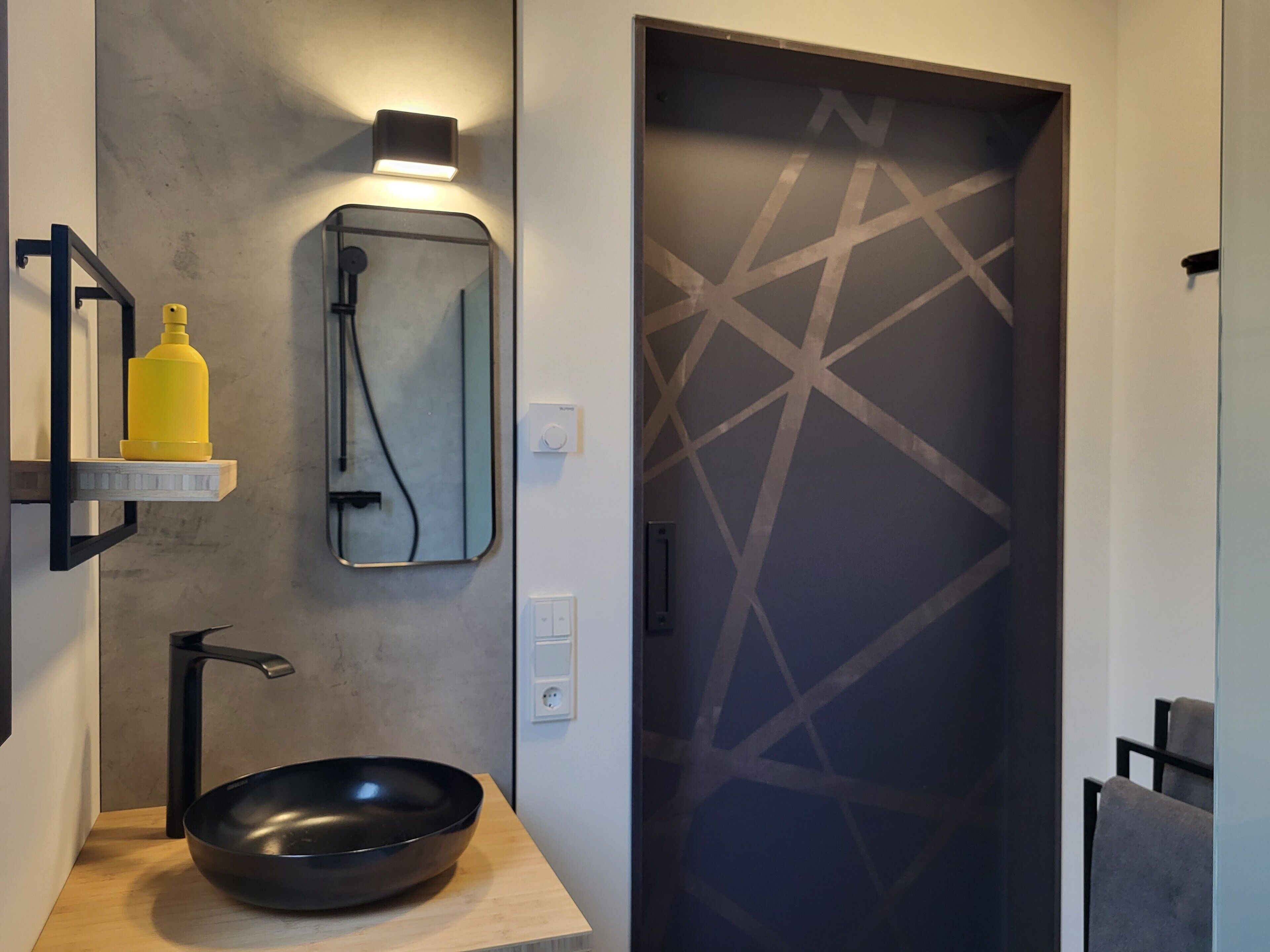 Un baño moderno con un lavabo negro, un espejo, una lámpara empotrada en la pared y una puerta con motivo geométrico.
