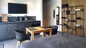 Ein modernes Hotelzimmer mit einer Küchenzeile, einem Essbereich mit Tisch und Stühlen, einem Bett und einer Wandablage mit Gläsern und Tassen.