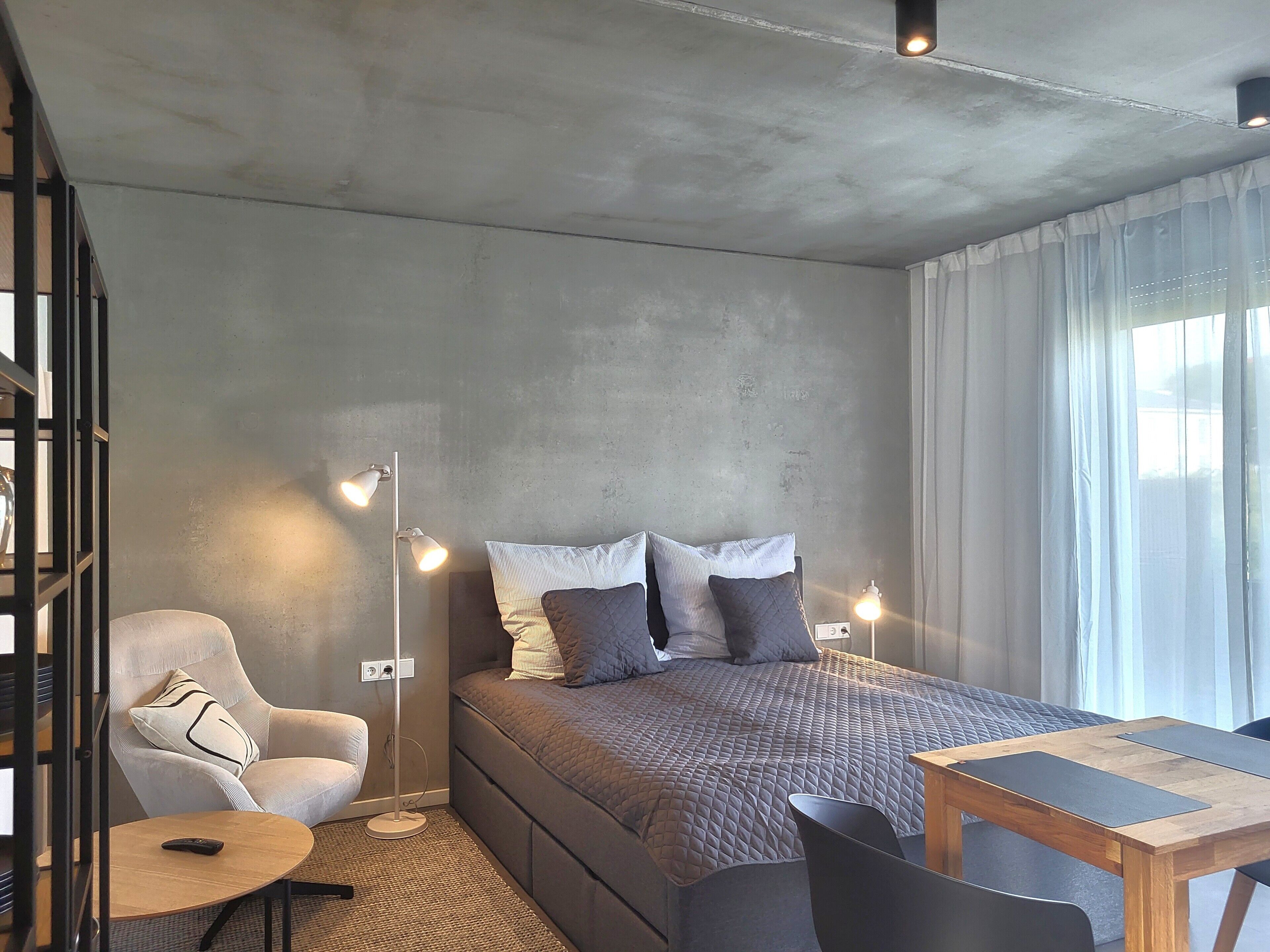 Ein modernes Schlafzimmer mit Bett, Nachttischlampen, einem Sessel, einem kleinen Tisch und einem Bücherregal.