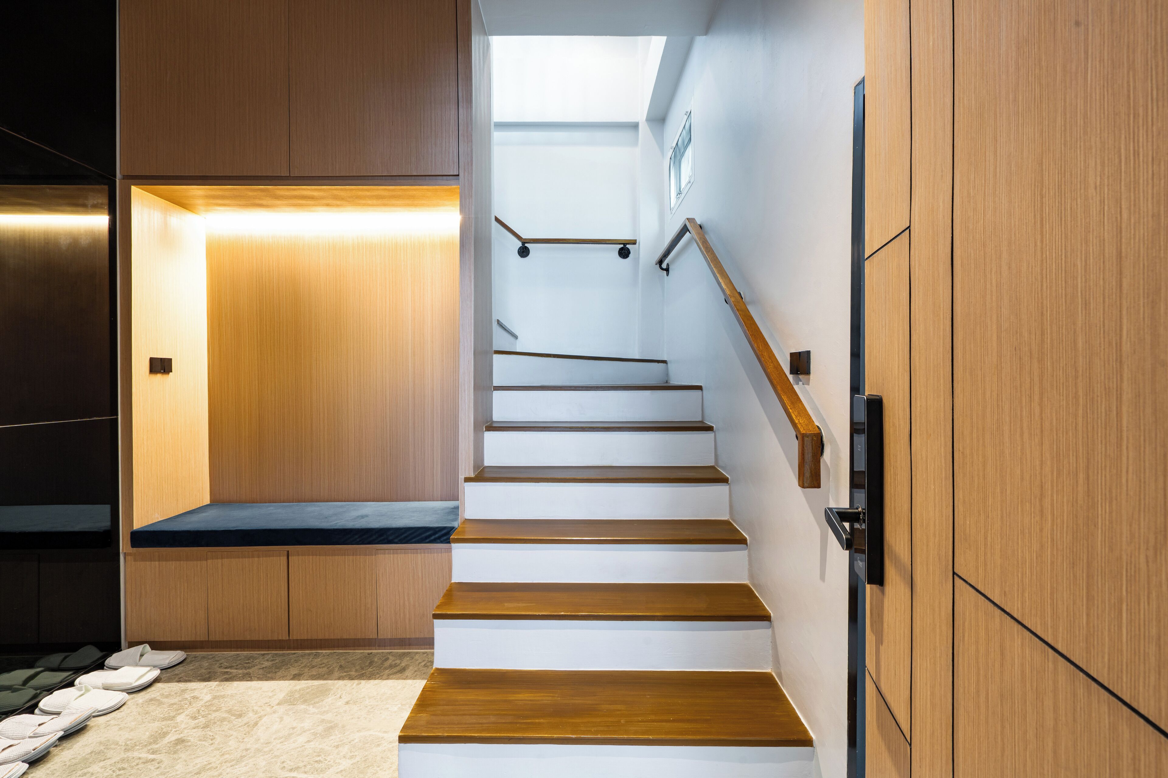 A modern staircase with wooden steps and a black handrail, leading to a landing with a bench and a wall-mounted light fixture.