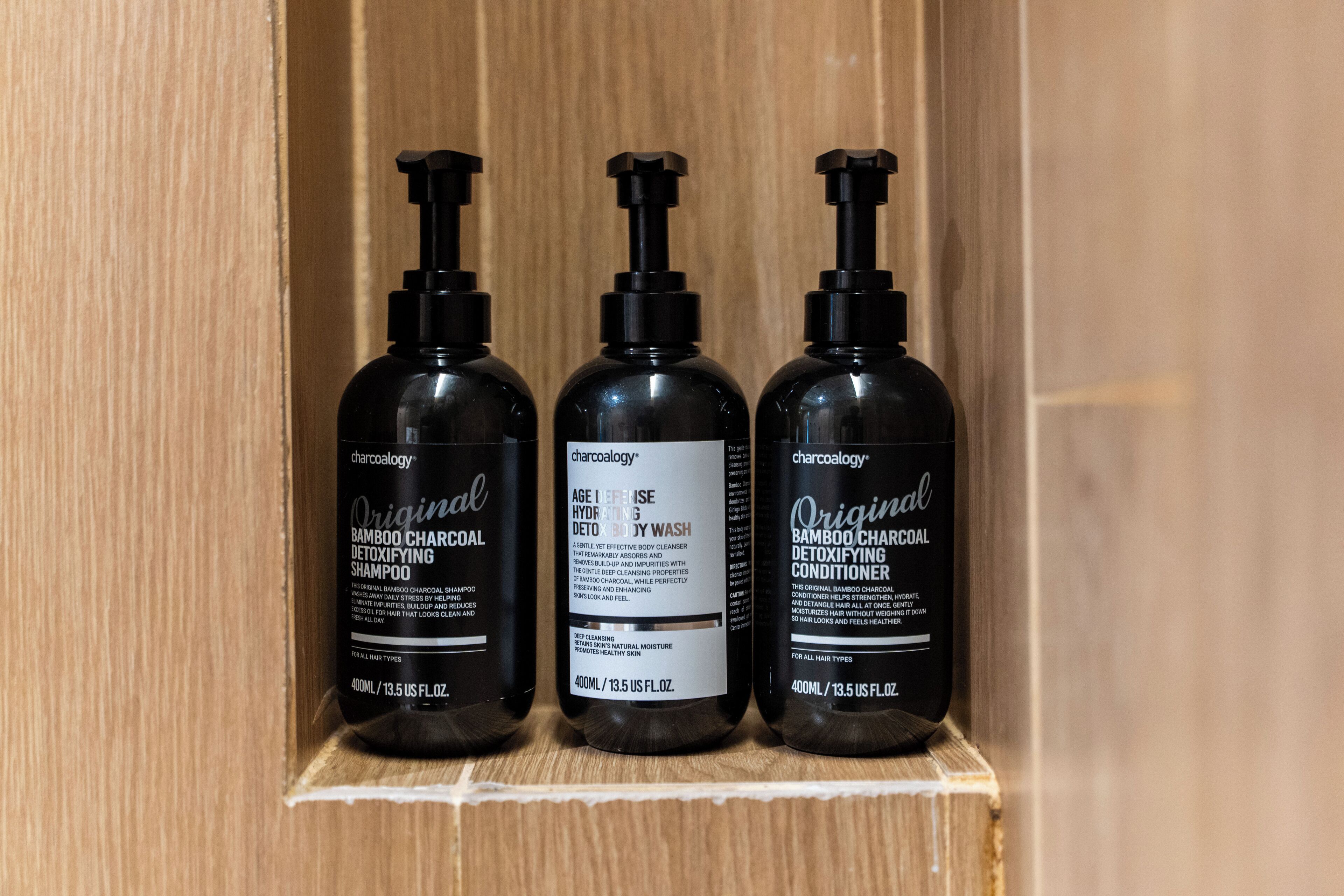 Tre flasker Charcoalology Original Bamboo Charcoal Detoxifying Shampoo og Conditioner på en træhylde.