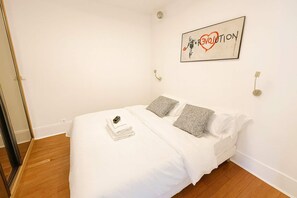 A neatly made bed with white linens and two grey pillows, a small stack of folded towels, and a framed artwork on the wall.