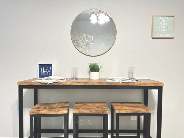 Un coin repas avec une table en bois, trois tabourets assortis, une plante en pot et un panneau encadré accroché au mur.