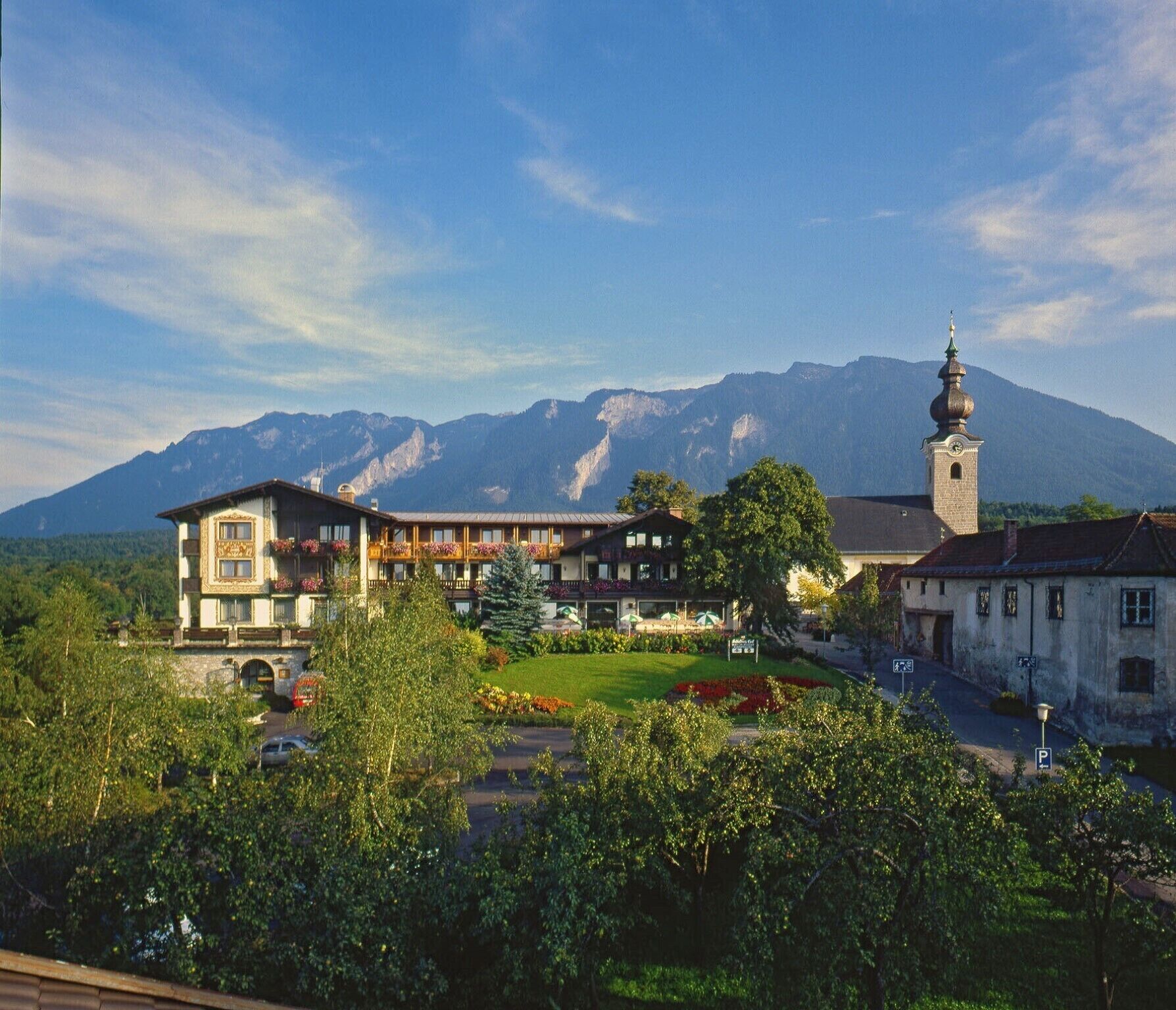 Un hôtel situé au pied d’une montagne, avec un clocher d’église imposant et une végétation luxuriante.
