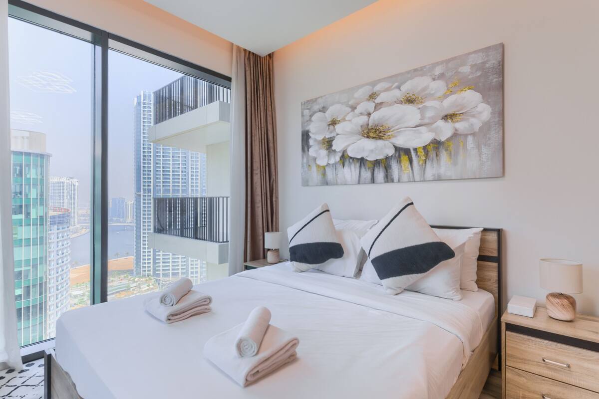 A hotel room with a large bed, a nightstand, a lamp, and a painting of flowers.