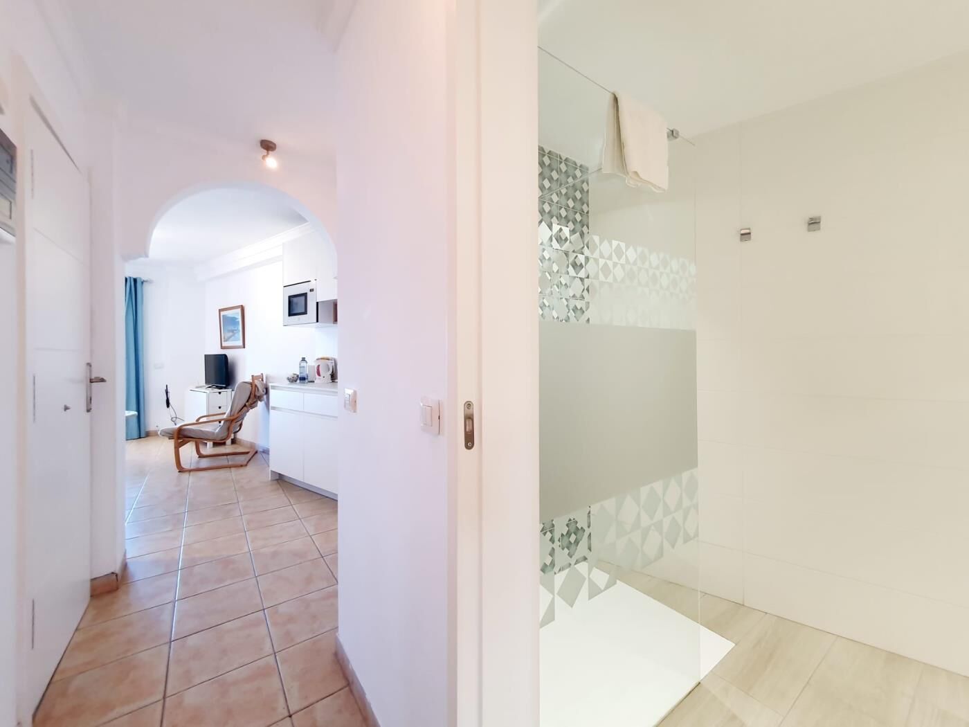 Una habitación de hotel moderna con suelo de baldosas, cabina de ducha de vidrio y una pequeña zona de estar con una silla y una mesa.