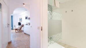 A modern hotel room with a tiled floor, a glass shower enclosure, and a small seating area with a chair and a table.