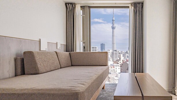 A modern hotel room with a sofa, a wooden side table, and a large window offering a view of a cityscape and a tower.