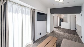 A modern hotel room with a large window, a flat-screen TV mounted on the wall, a wooden bench, and a carpeted floor.