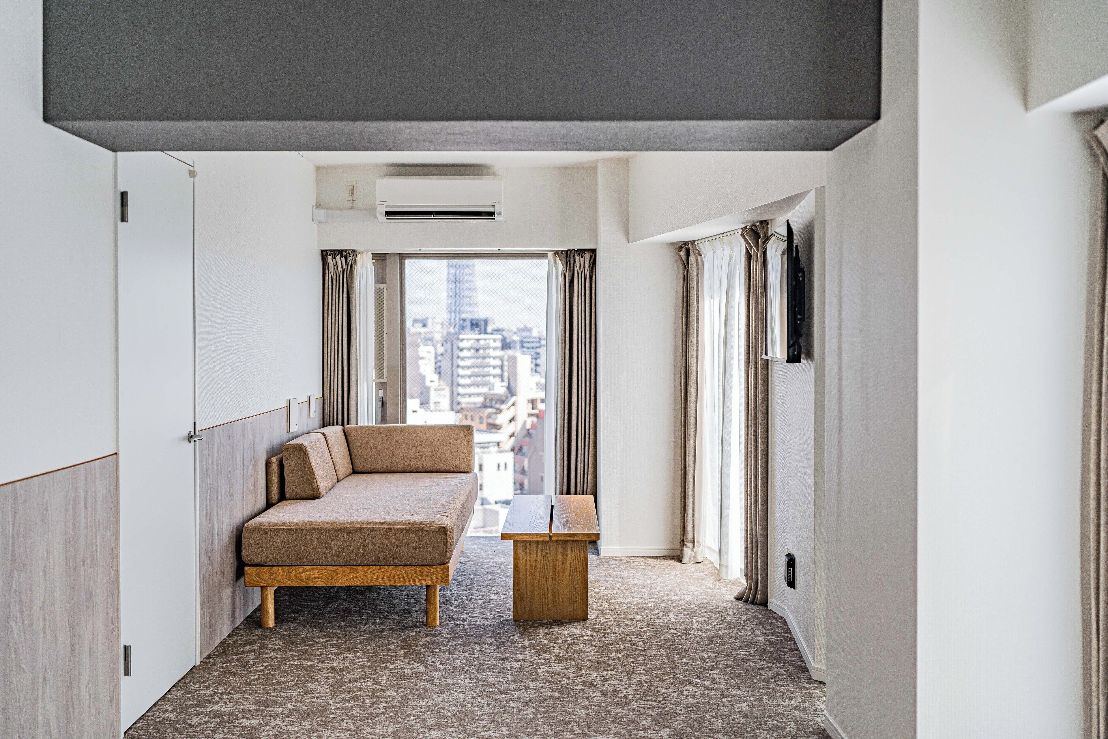 Ett modernt hotellrum med en soffa, ett litet träbord och utsikt över staden genom stora fönster.