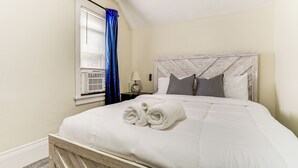 A neatly made bed with white bedding and rolled towels, a wooden headboard, a nightstand with a lamp, and a window with blue curtains. - 1 Mi to University: Spacious Home in Minneapolis! (Minneapolis)