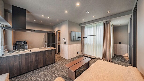 A modern hotel room with a kitchenette, a large bed, a bench, a TV, and a sliding glass door to a balcony.