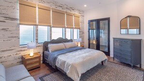 Un dormitorio con cama, mesitas de noche, cómoda, espejo y ventanas amplias con persianas.