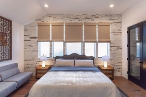 A bedroom with a large bed, two bedside tables with lamps, a grey sofa, and a window with blinds.