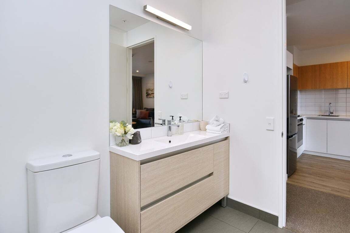 A modern bathroom with a white toilet, a large mirror, a wooden vanity with a sink, and a visible shower area.