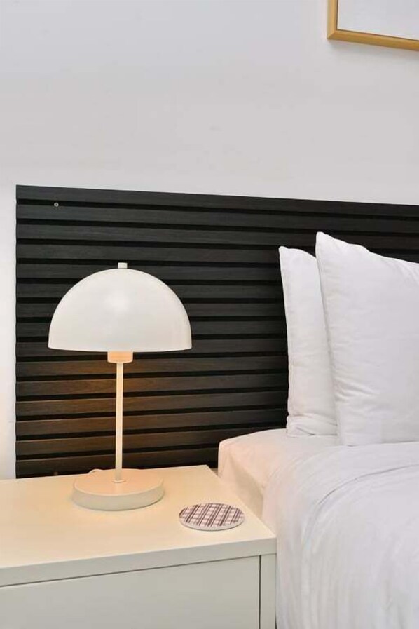 Una cama con ropa de cama blanca, una mesita de noche blanca con una lámpara y un cabecero de madera oscura.