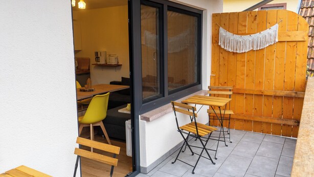 A patio area with wooden tables and chairs, a yellow chair, and a wooden fence with a fringe decoration.