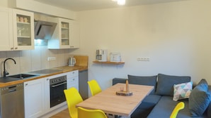 A compact kitchen with white cabinets, a dishwasher, and a stove. There is a wooden table with a yellow chair and a gray sofa with pillows.