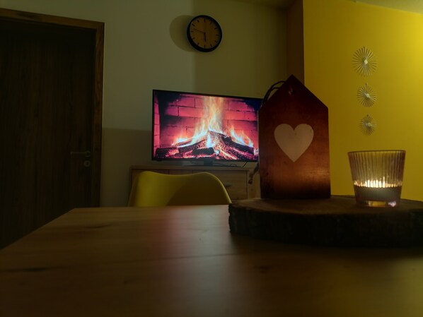 Una habitación con una mesa de madera, un televisor que muestra una chimenea, una decoración con forma de corazón y un candelabro.