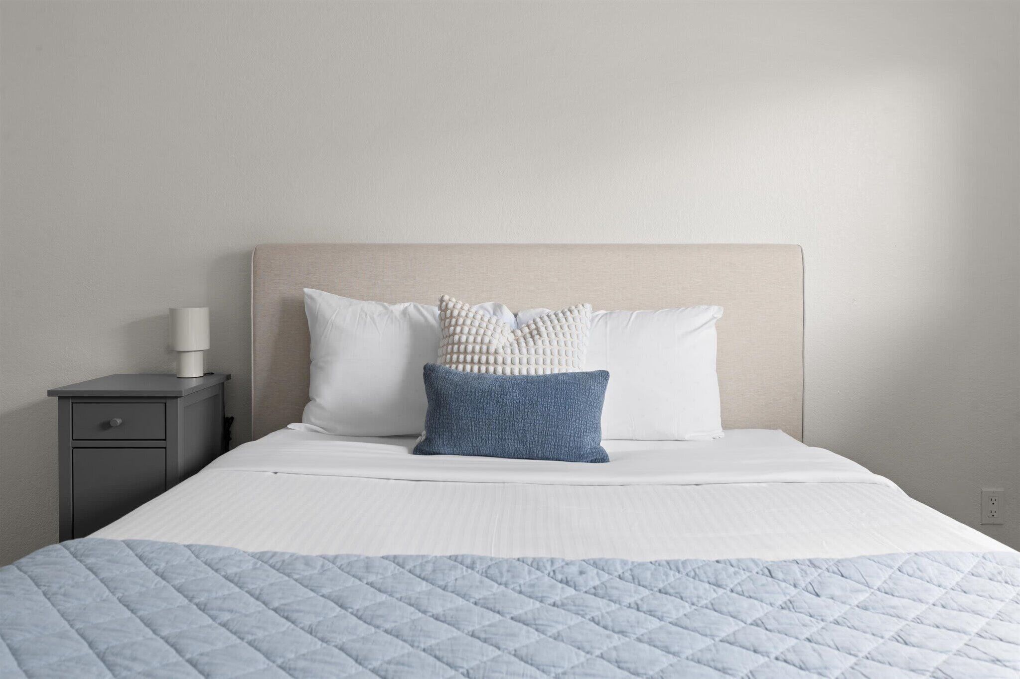 A neatly made bed with a quilted blue blanket, two pillows, and a bedside lamp on a gray nightstand.