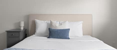 Una cama bien hecha con un edredón azul acolchado, dos almohadas y una lámpara de noche en una mesita de noche gris.