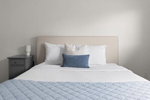 A neatly made bed with a quilted blue blanket, two pillows, and a bedside lamp on a gray nightstand. - The Sun Club | Private Pool • BBQ • Outdoor Dining (Mesa)