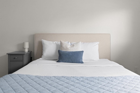 A neatly made bed with a quilted blue blanket, two pillows, and a bedside lamp on a gray nightstand.
