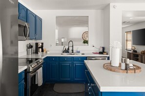 A modern kitchen with blue cabinets, stainless steel appliances, a white countertop, and a wooden cutting board with utensils.
