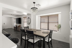 A dining area with a wooden table, four chairs, and a chandelier. There are two windows with blinds, a television on the wall, and a potted plant on the windowsill.