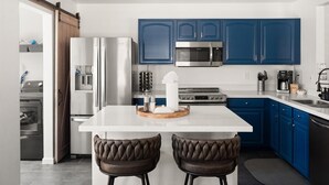 A modern kitchen with blue cabinets, stainless steel appliances, a central island with stools, and a sliding barn door. - The Sun Club | Private Pool • BBQ • Outdoor Dining (Mesa)